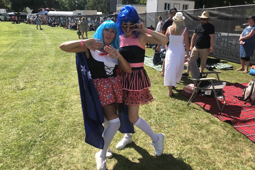 Two women stand on grass posing in brightly coloured outfits and blue wigs with a crowds behind them on a sunny day.