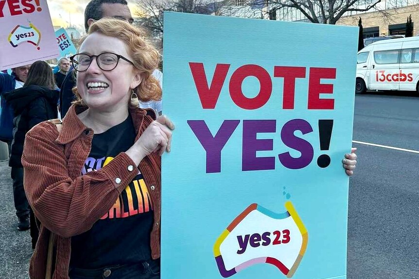 A young woman with glasses holds a sign reading: Vote Yes