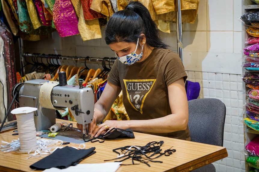 A woman sits while using a sewing machine to make a fabric mask.