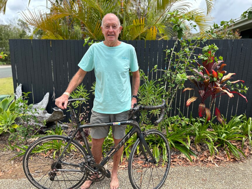 A fit but older man stands outside in his garden with a bicycle. 