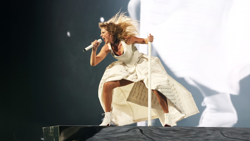 Taylor Swift singing passionately into a microphone, in a white dress shaking her head