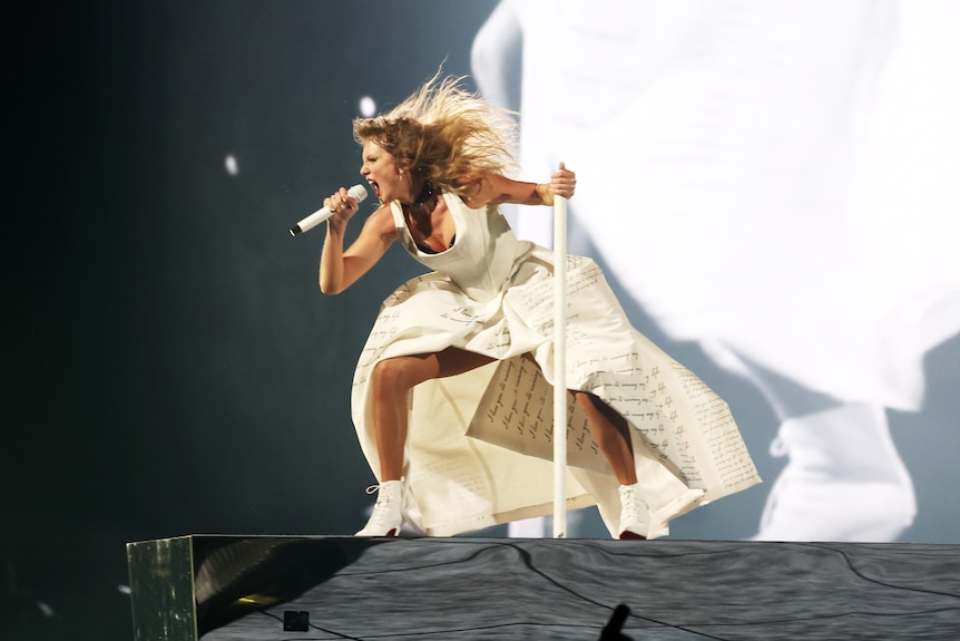 Taylor Swift singing passionately into a microphone, in a white dress shaking her head