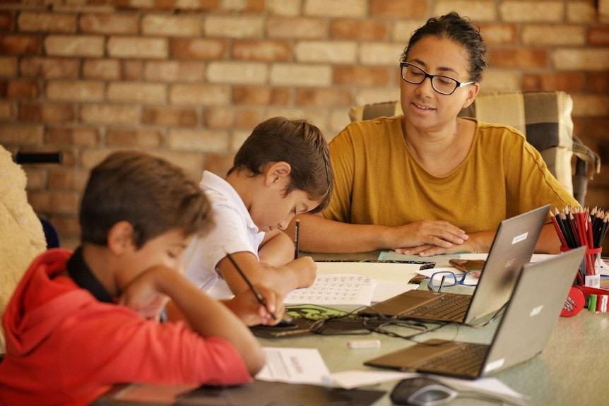 A woman sits with her two boys watching them writing on worksheets with their laptops open in front of them
