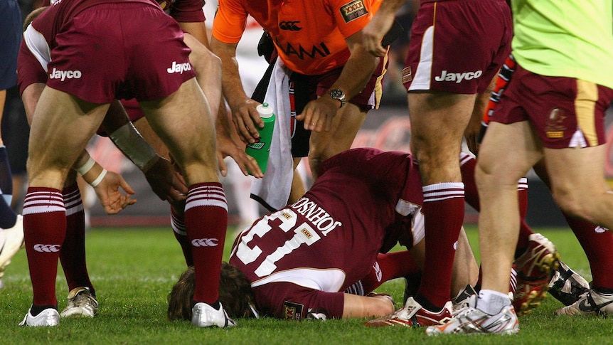 Queensland Maroons players and trainers gather around Dallas Johnson, who is face down in the ground during an Origin match.