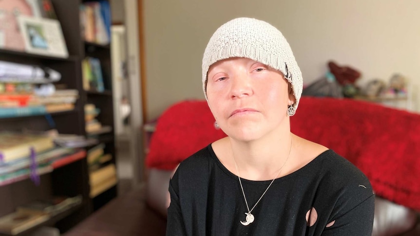 A woman wearing a beanie and a black shirt sits in front of a book shelf looking at the camera.