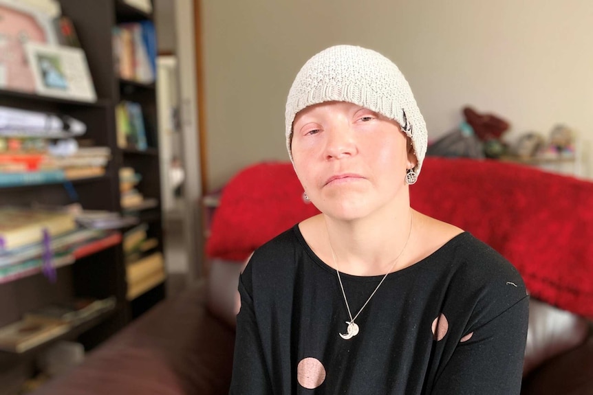 A woman wearing a beanie and a black shirt sits in front of a book shelf looking at the camera.