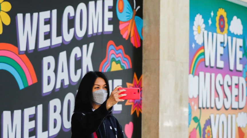 A woman takes a selfie in front of a sign reading 'welcome back Melbourne'.