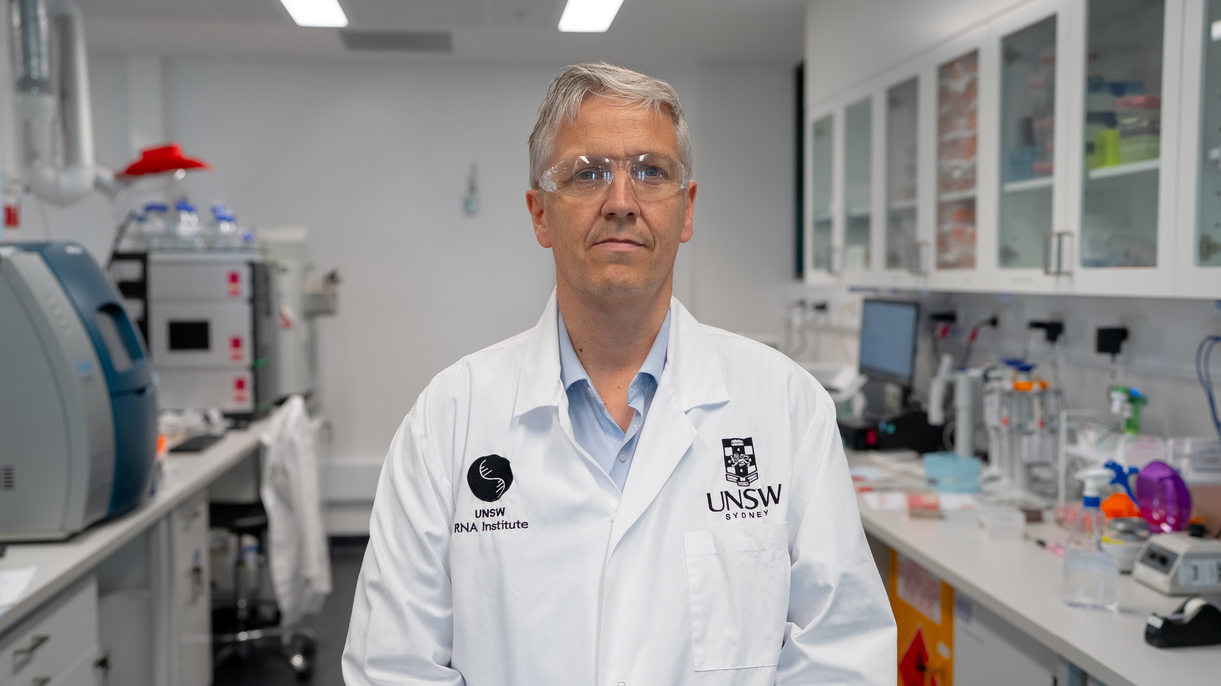 A grey haired man in a lab coat wearing safety glasses.