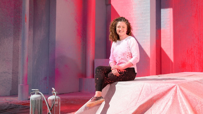 A smiling woman with long, curly hair sits in an art gallery.