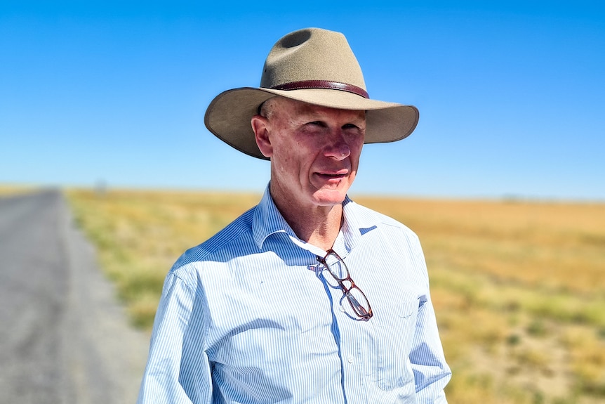 A man in a broad-brimmed hat stands in bright sunlight flooding a dusty paddock.