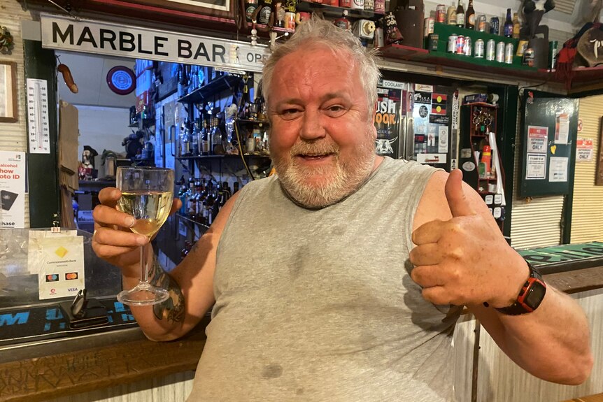A man in stained grey singlet, with grey hair and beard, smiles at camera, holding a glass of wine and giving thumbs up