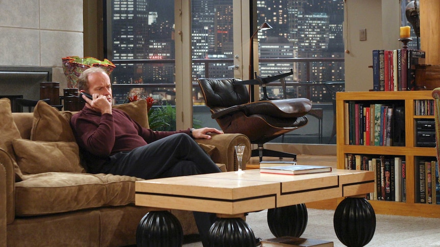 On a rainy night, Frasier sits on a brown couch in his apartment while holding a wireless landline phone up to his ear.