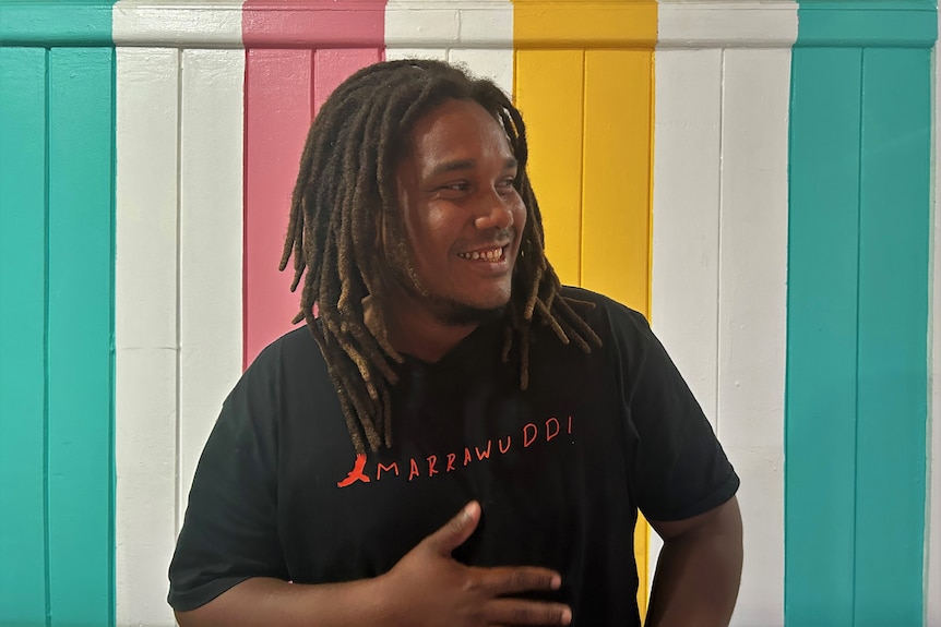 A solomon islands man with long dreadlock hair smiles in front of a rainbow coloured wall 
