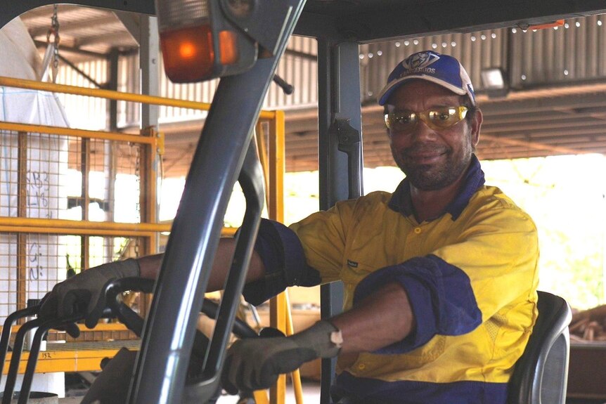 A man wearing hi-vis and goggles smiles as he operates a forklift in a large shed.