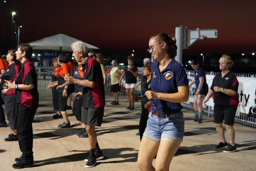 A group of people line dancing in an outdoors area at dusk, with a young woman wearing a T-shirt and denim shorts in the front.