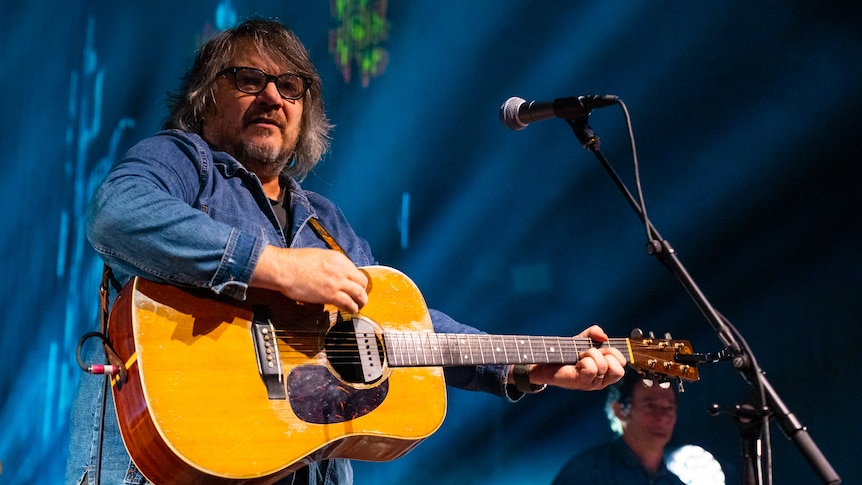 A man wearing a blue shirt and glasses plays an acoustic guitar on stage