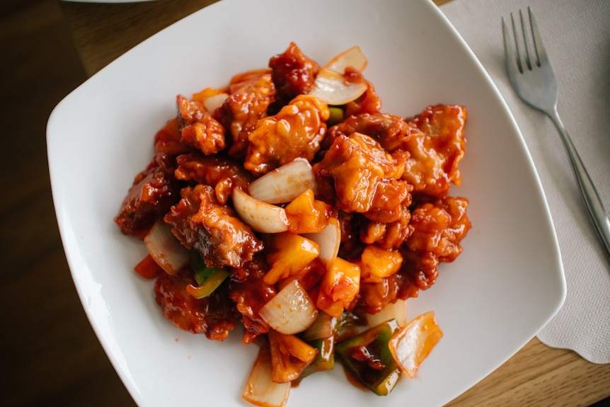 Small chunks of pork and vegetables cooked together in a sticky red sauce sit on a white plate.