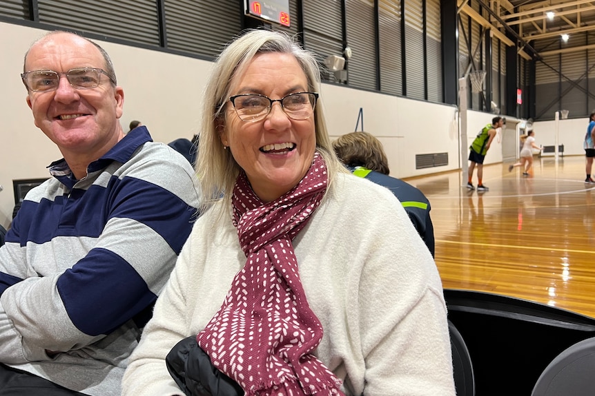 Michelle wears a white jumper purple scarf and glasses she sits next to other parents watching the game