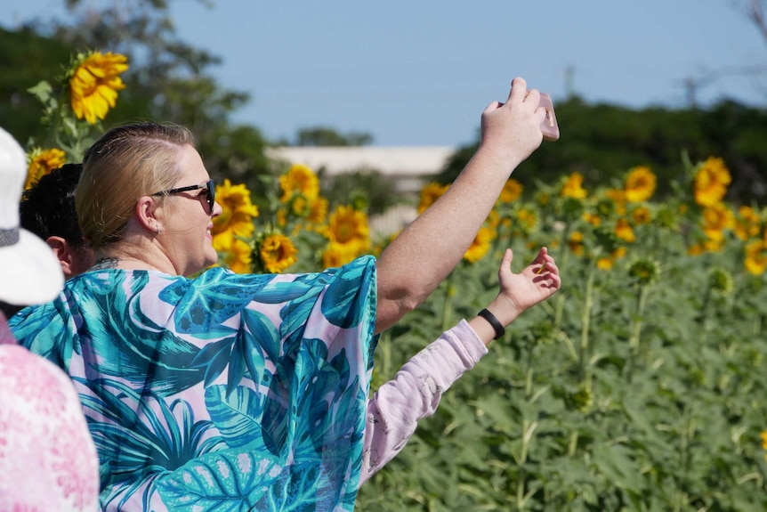 A woman holds up her phone for a selfie in front of a sunflower crop