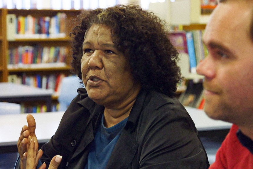 An Indigenous woman sitting in a library gesticulates while she talks.