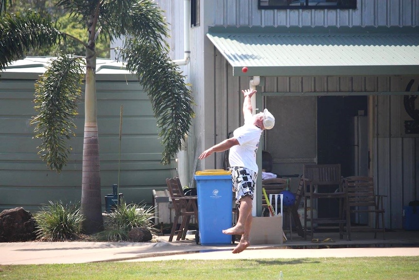 A man wearing a white shirt and cap and floral board shirts jumping to catch a red cricket ball.