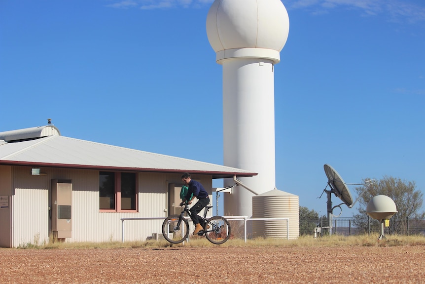 The white weather station and the distant figure of a man on his bike, wears blue shirt, black jeans, blue sky, red earth.