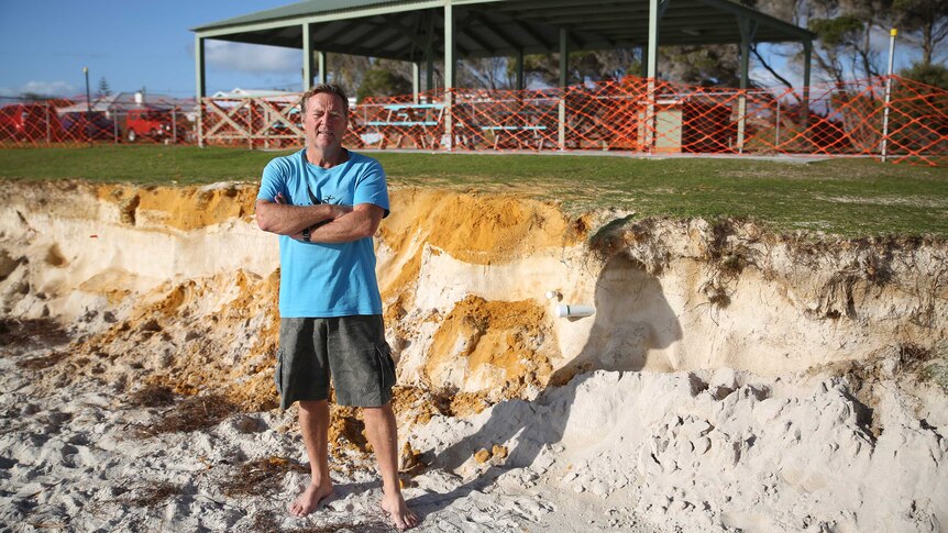 A man in a blue t-shirt and short stands in front of an eroded stretch of beach, next to a barbecue shelter.