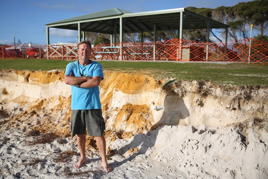 A man in a blue t-shirt and short stands in front of an eroded stretch of beach, next to a barbecue shelter.