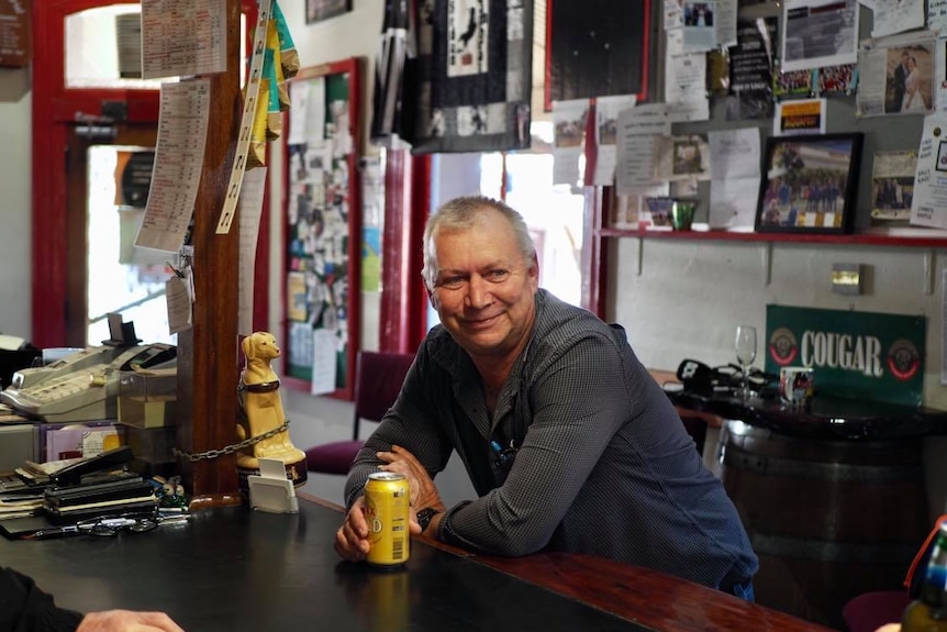 A middle aged man wearing a grey long sleeved shirt leans over a bar, holding a gold can of beer. He is smiling.