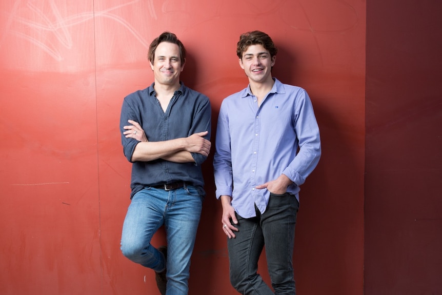 A 40-something man and a 20-something man stand next to each other, smiling proudly and leaning against a red wall