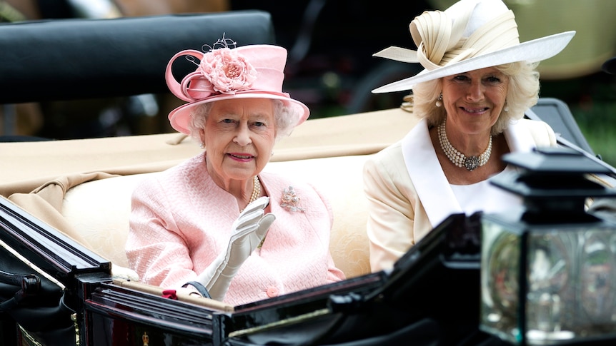 Queen Elizabeth and Camilla sit side by side in a carriage smiling and waving 