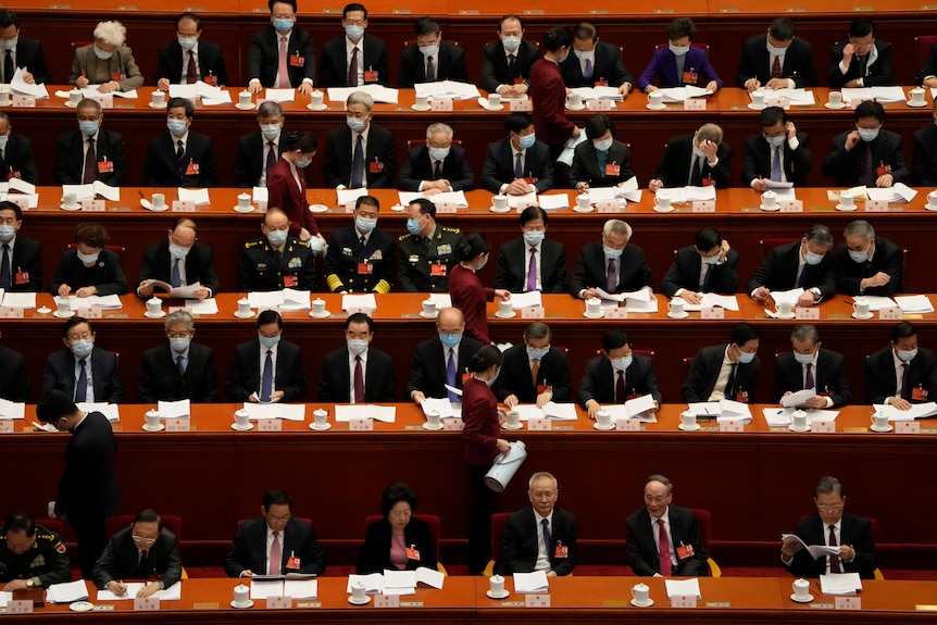 A group of men sitting in China's National People's Congress, as two women in red pour tea.