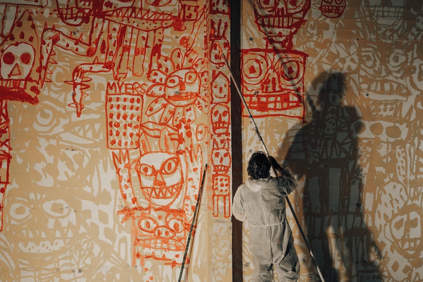 Ein Mann im Plastikanzug, der einen Pinsel an einer Stange hält, malt ein orangefarbenes Wandbild an eine Wand