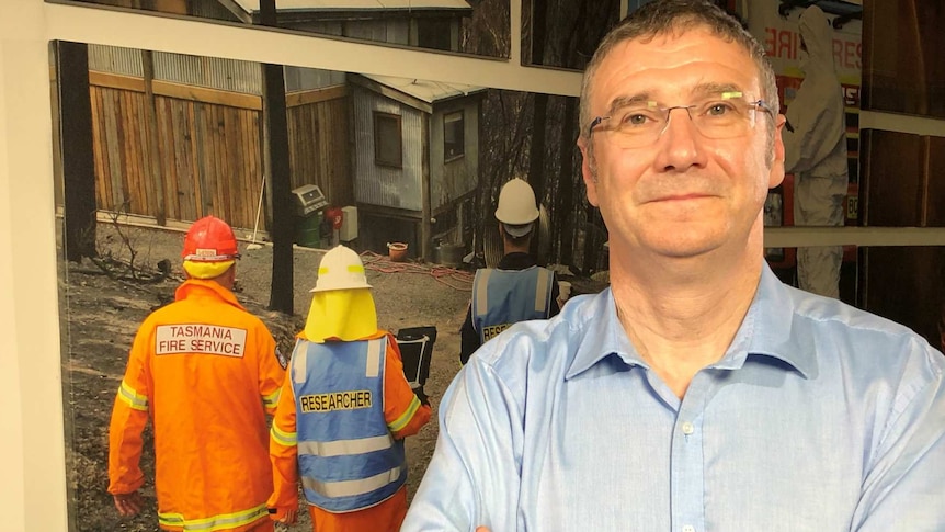 Richard Thornton, dressed in office attire, stands in front of an image of firefighters on a fire ground.