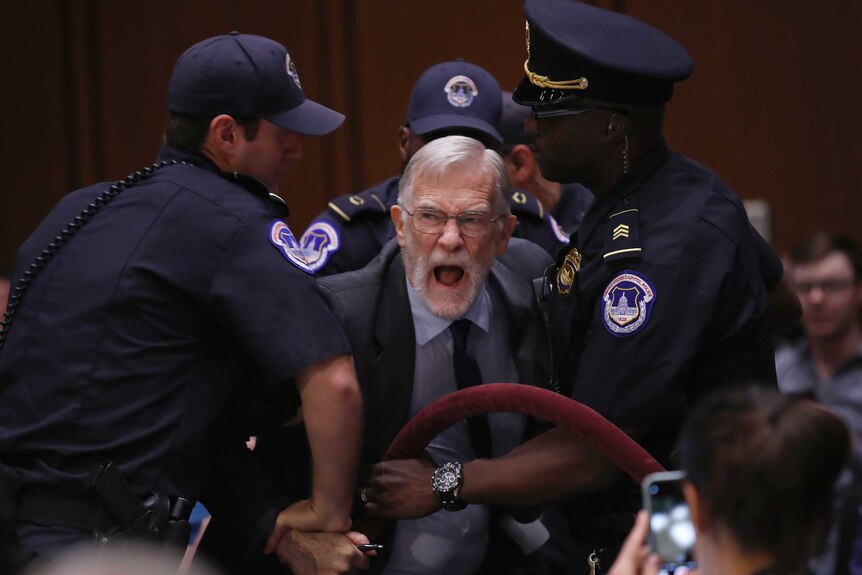A protester, dressed in a suit and yelling, is restrained and removed.