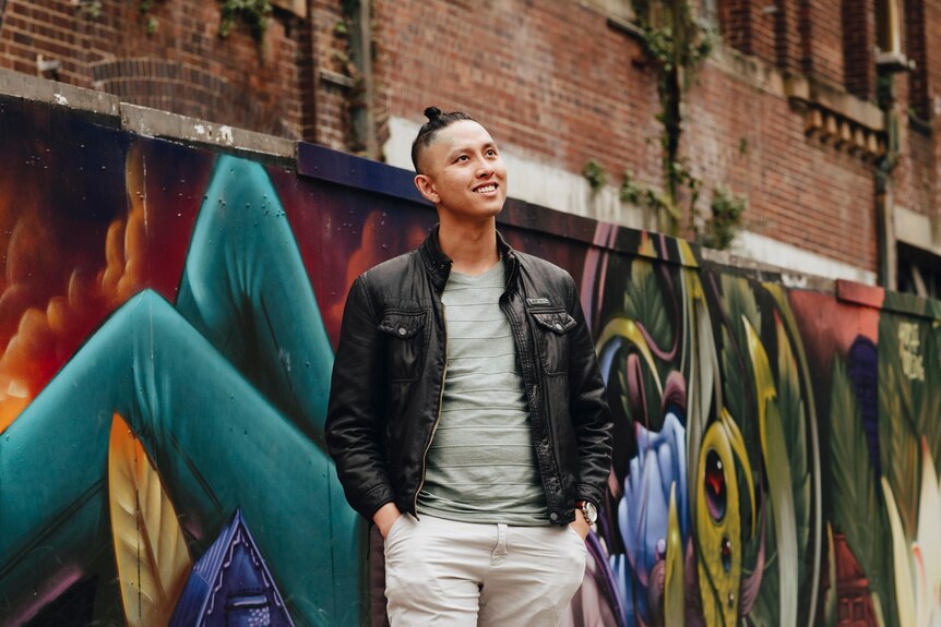 Mid-20s Vietnamese Australian man Peter Nguyen standing in front of graffiti and brick wall, looking up and smiling slightly. 