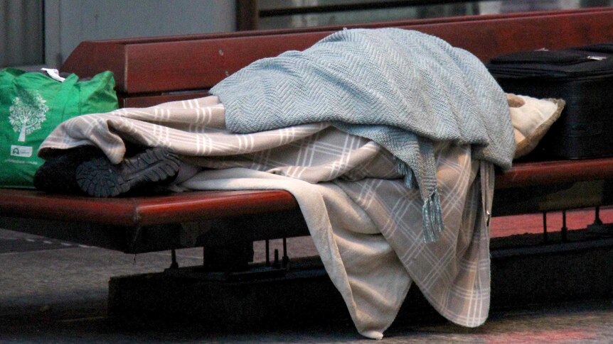 Wide shot of a homeless person sleeping on a bench.