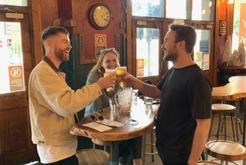 Two men in a pub clink filled schooner glasses together as a woman at their table watches and smiles.