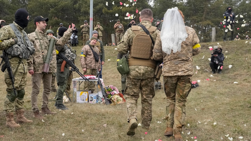 A couple wearing military uniforms walk through a field, the woman wearing a veil, as other service people throw rose petals
