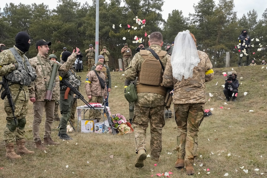 A couple wearing military uniforms walk through a field, the woman wearing a veil, as other service people throw rose petals