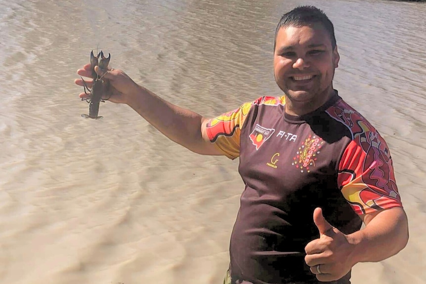 A man holds a yabby with a murky brown river behind