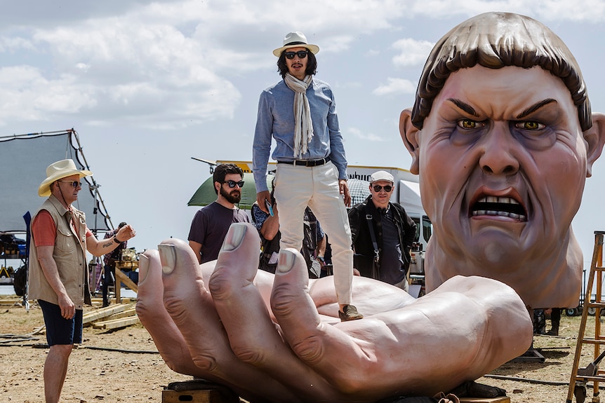 Colour still of Adam Driver standing on giant hand prop on film set as Toby Grisoni in 2018 film The Man Who Killed Don Quixote.