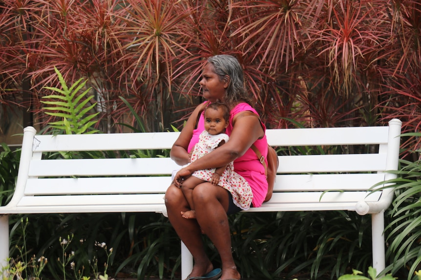 Darlene Anne Wauchope sits on a white bench in front of plants with a child in her lap.