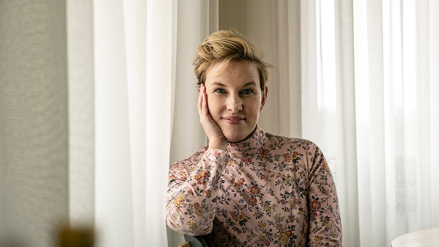 Actress with short blonde hair sits at table with white curtains behind, wearing floral top, and resting chin on hand.