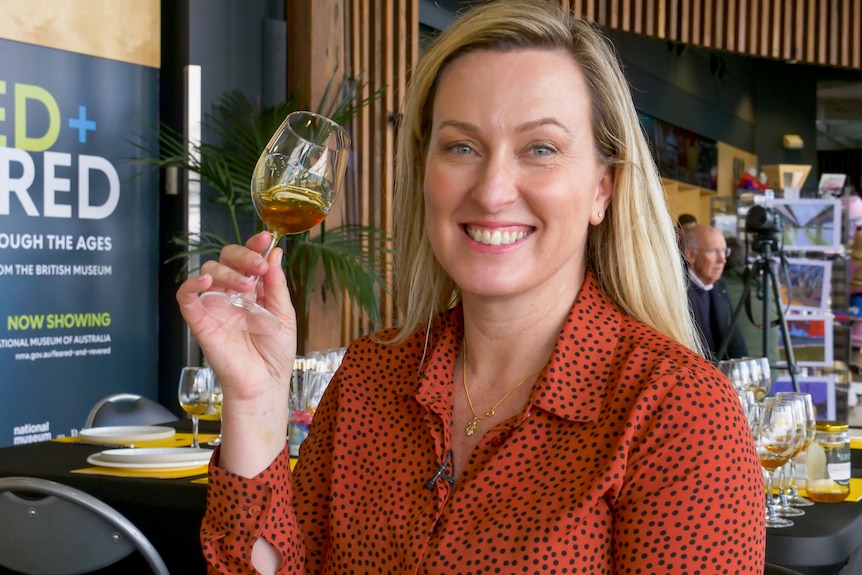 Woman with blonde hair wearing a burnt orange top with black spots, holding a wine glass containing honey. 