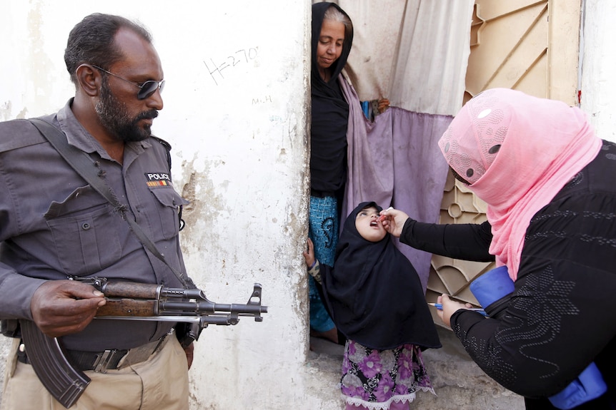 A policeman stands guard with a gun as a child is given the polio vaccine in Pakistan,