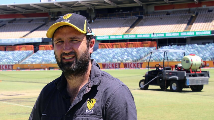 A man in a WACA cricket cap stands on a sporting arena ground.