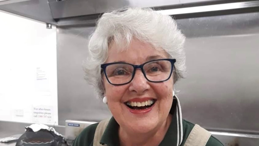 An elderly woman smiles broadly. She is wearing an apron and appears to be in a commercial kitchen.