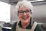 An elderly woman smiles broadly. She is wearing an apron and appears to be in a commercial kitchen.