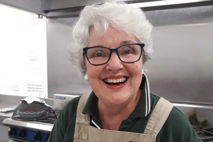 An elderly woman smiles broadly. She is wearing an apron and appears to be in a commercial kitchen.